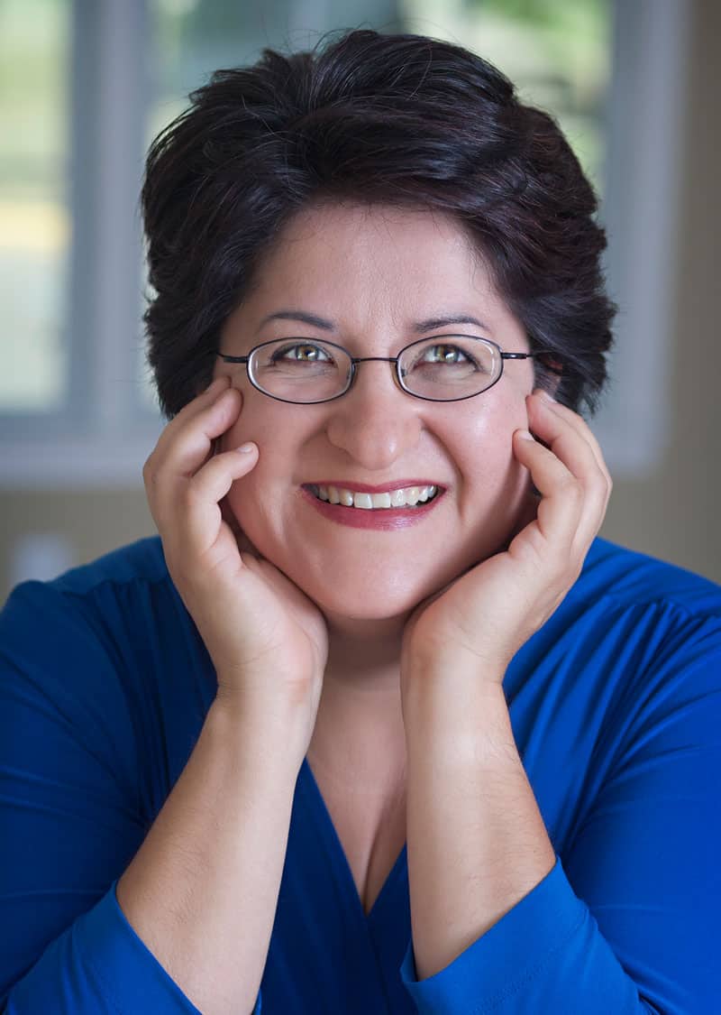 Valerie M. Trujillo in a blue shirt and glasses with her hands on her cheeks