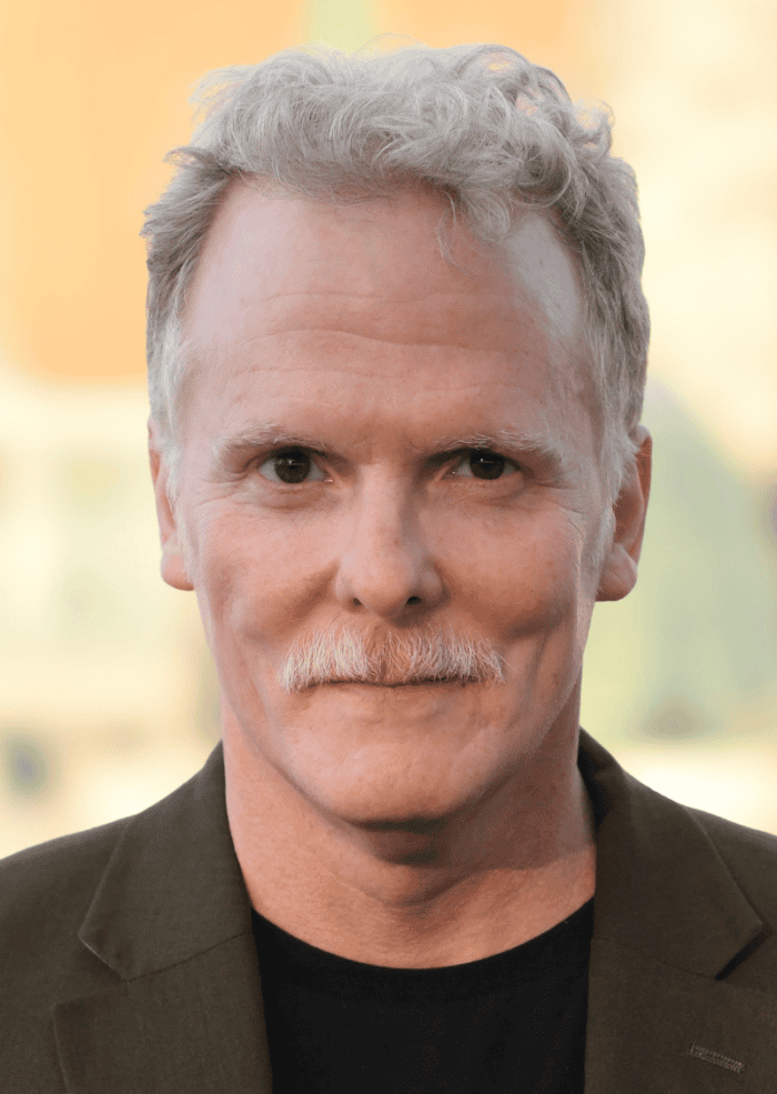 Daniel Pearce headshot. He has salt and pepper colored hair with a mustache. He is wearing a green suit jacket and a black shirt. 