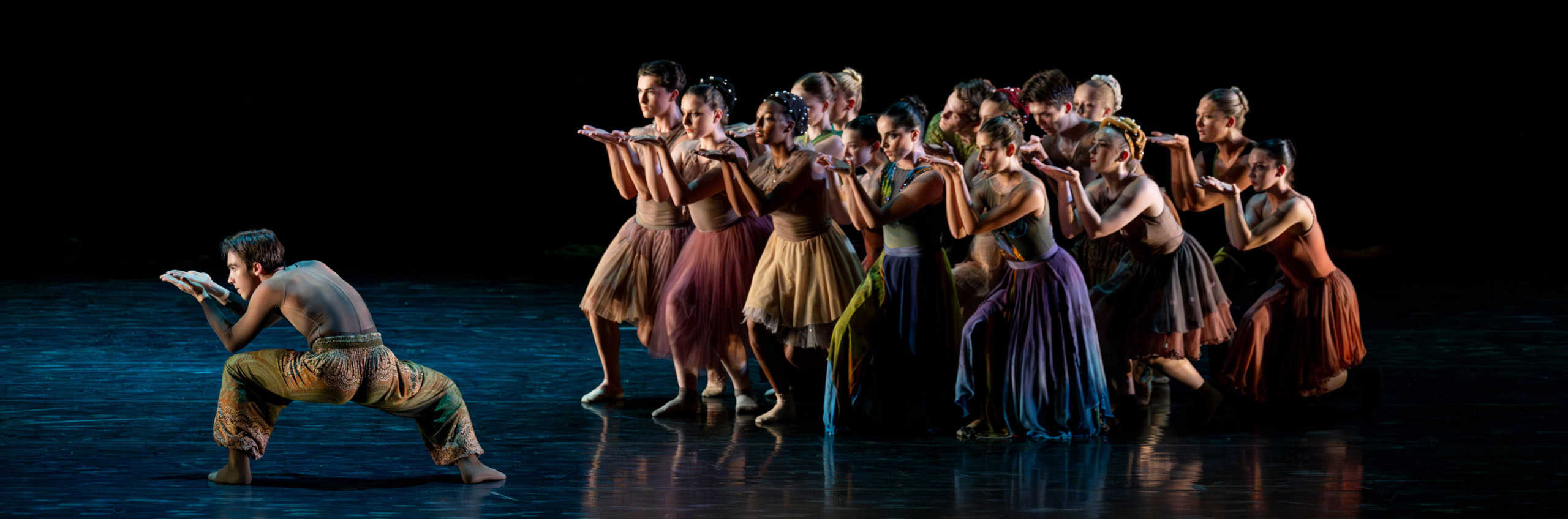 A male dancer crouching low with his palms raised and a group of dancers approaching behind him with their palms up