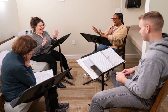 Four people rehearsing a play, reading from music stands